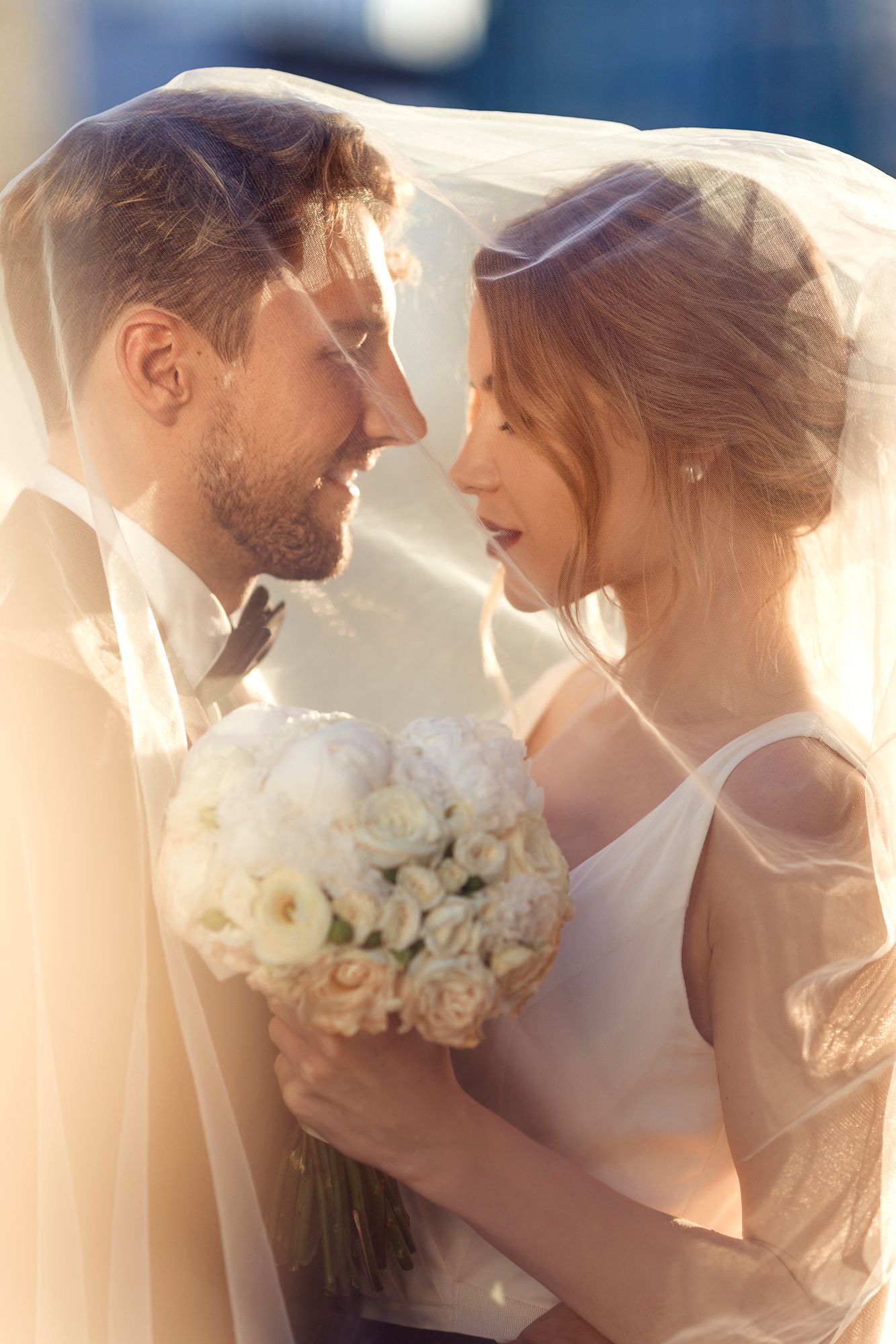 Romantic wedding portrait of couple sharing tender moment under a flowing veil at sunset.