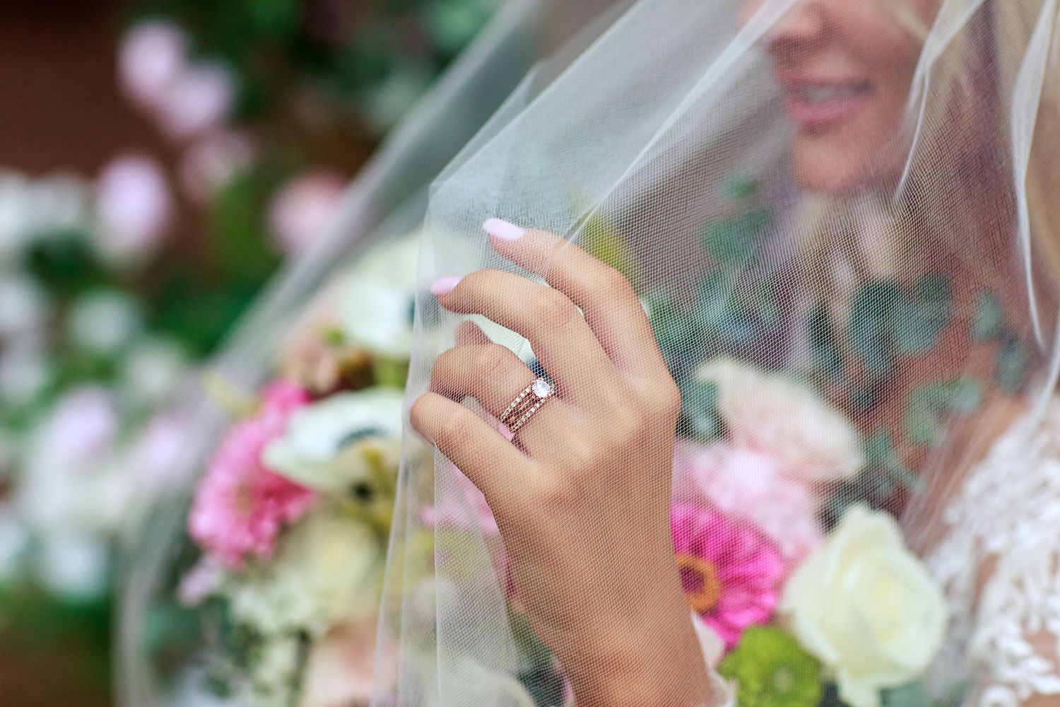 Close up of diamond engagement ring through bridal veil with flowers in background.