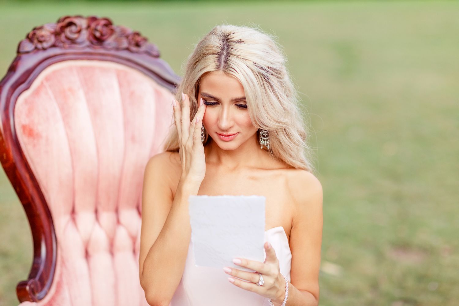 Woman in white dress seated on pink vintage chair outdoors reading letter.