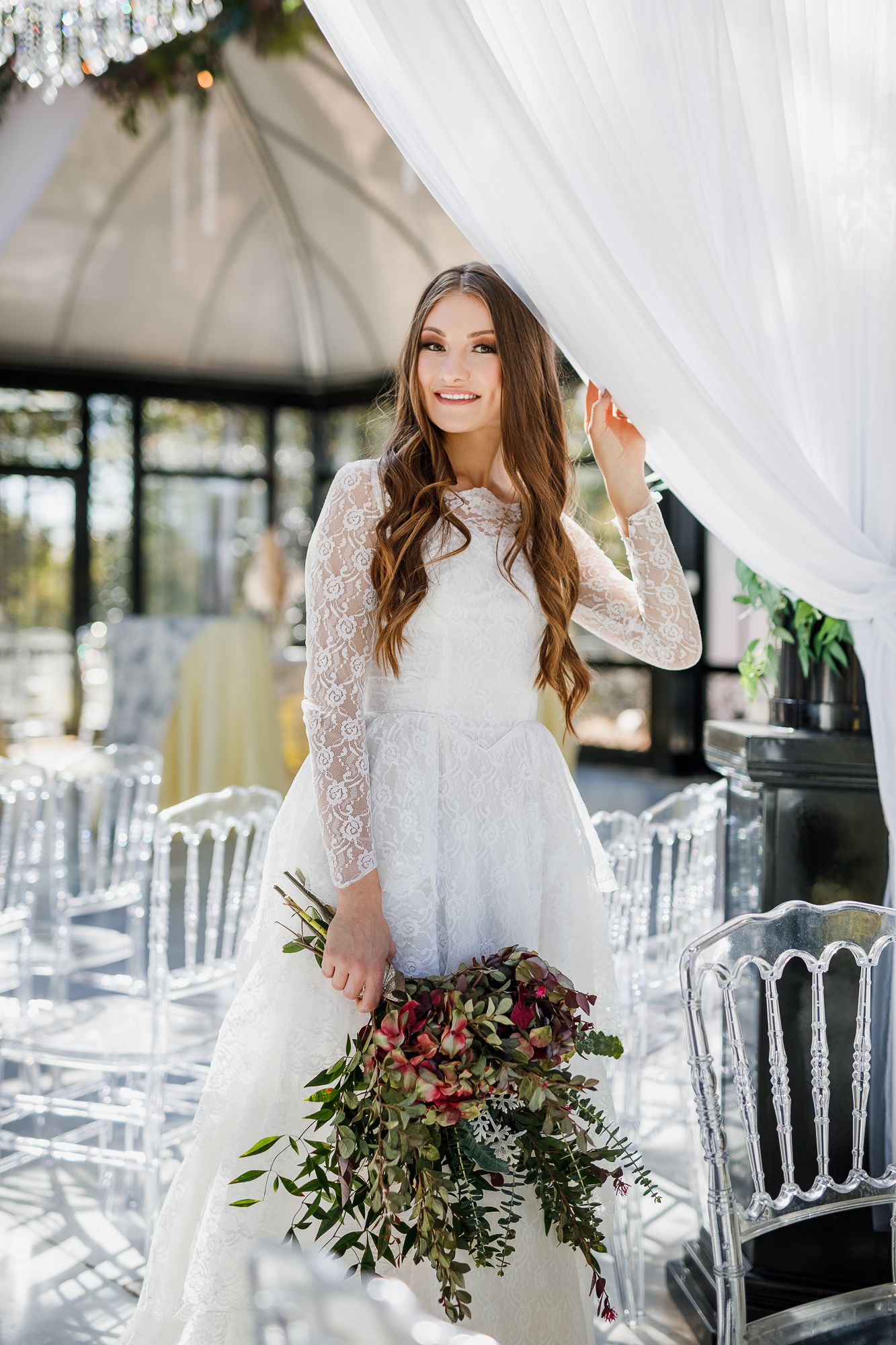 Bride in long-sleeve lace wedding gown holding cascading bouquet in elegant outdoor venue setting.