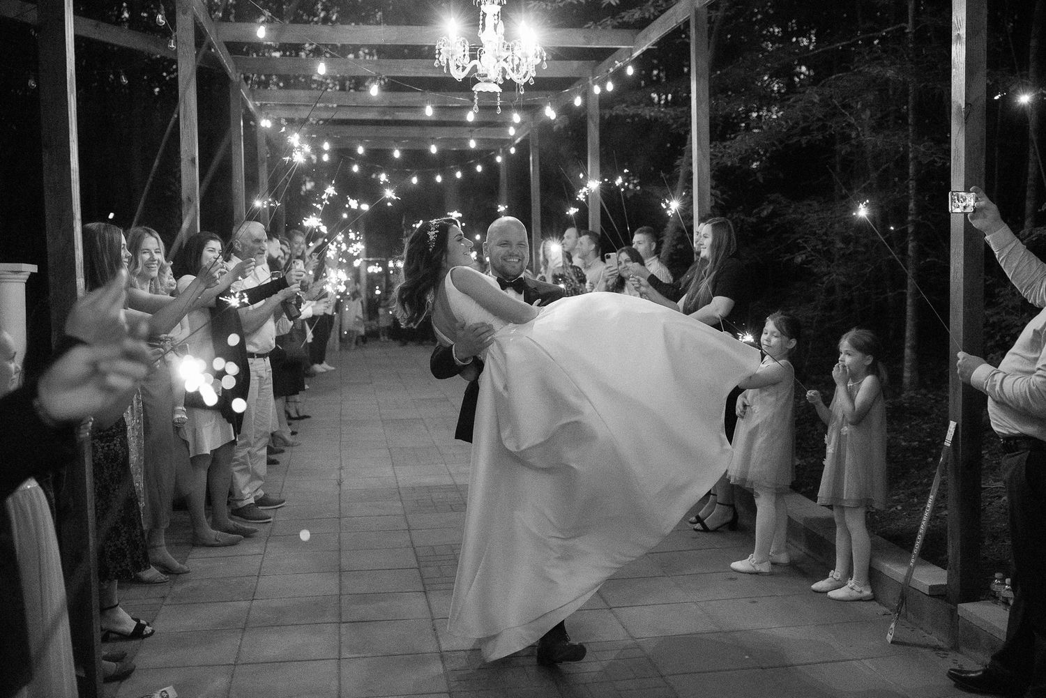 Black and white wedding exit photo with sparklers lighting pathway for dancing newlyweds.