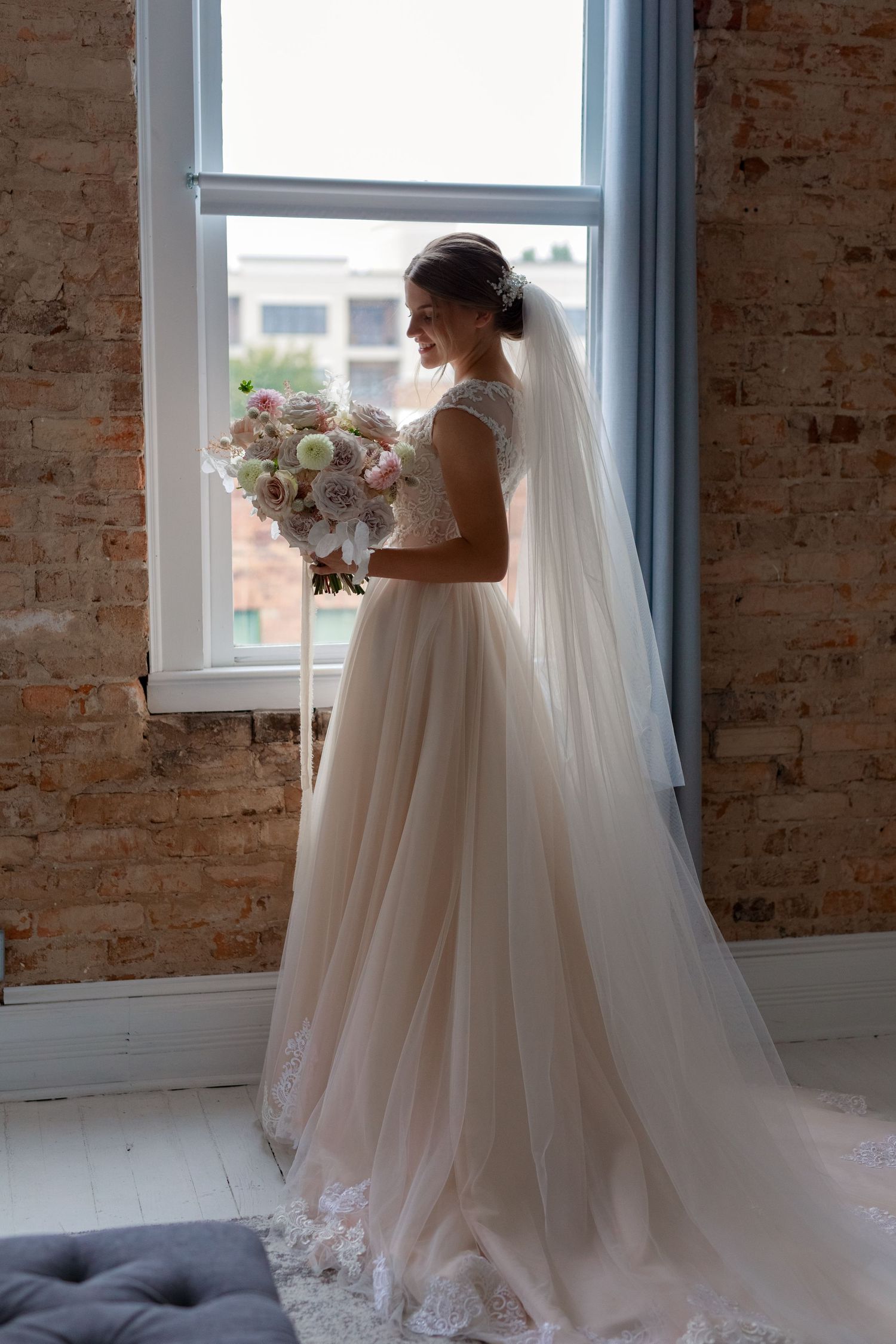 A bride in a flowing white gown stands by a window holding a pink and white bouquet.