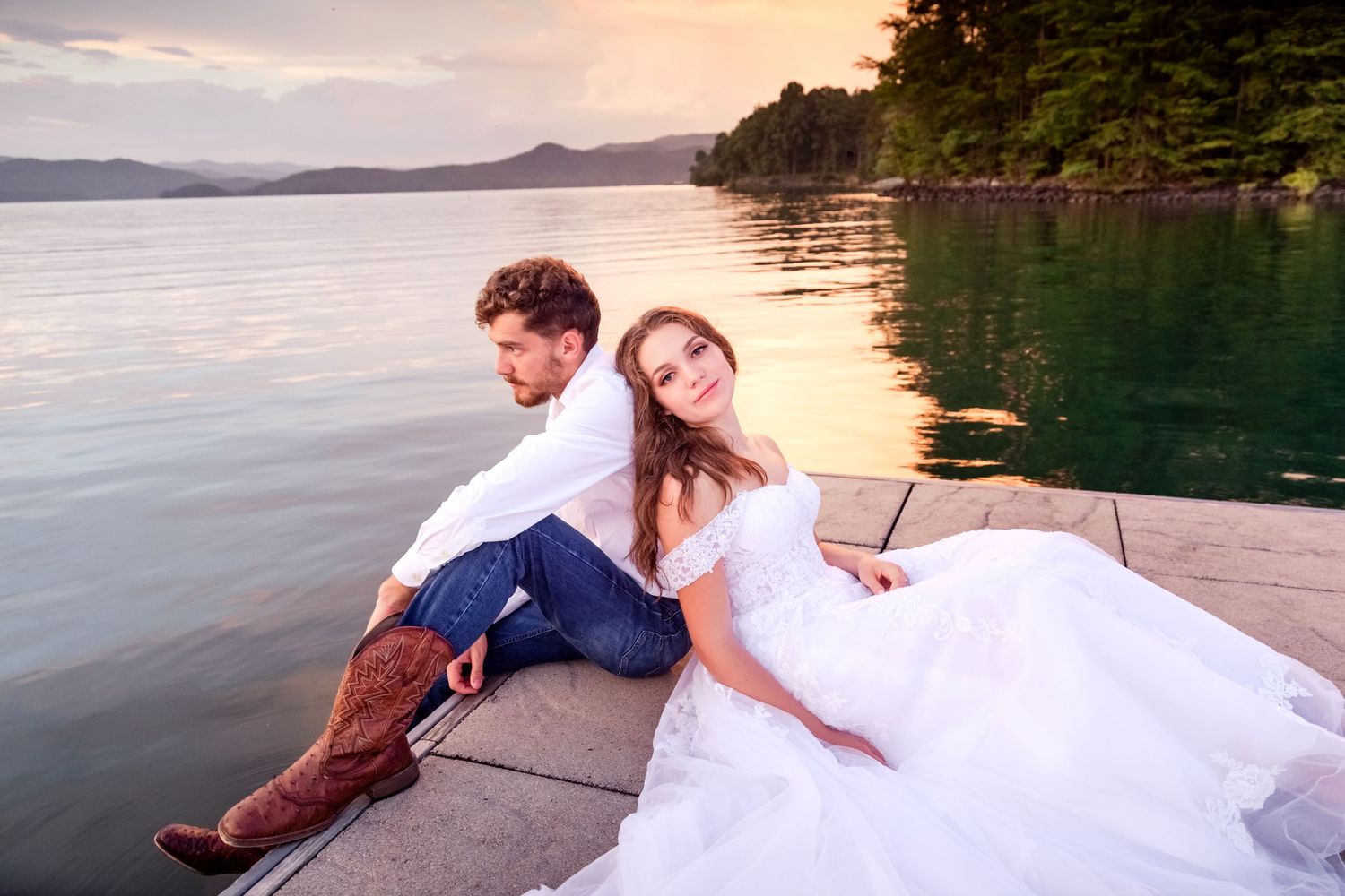Couple relaxing on a dock by a serene lake at sunset in wedding attire.