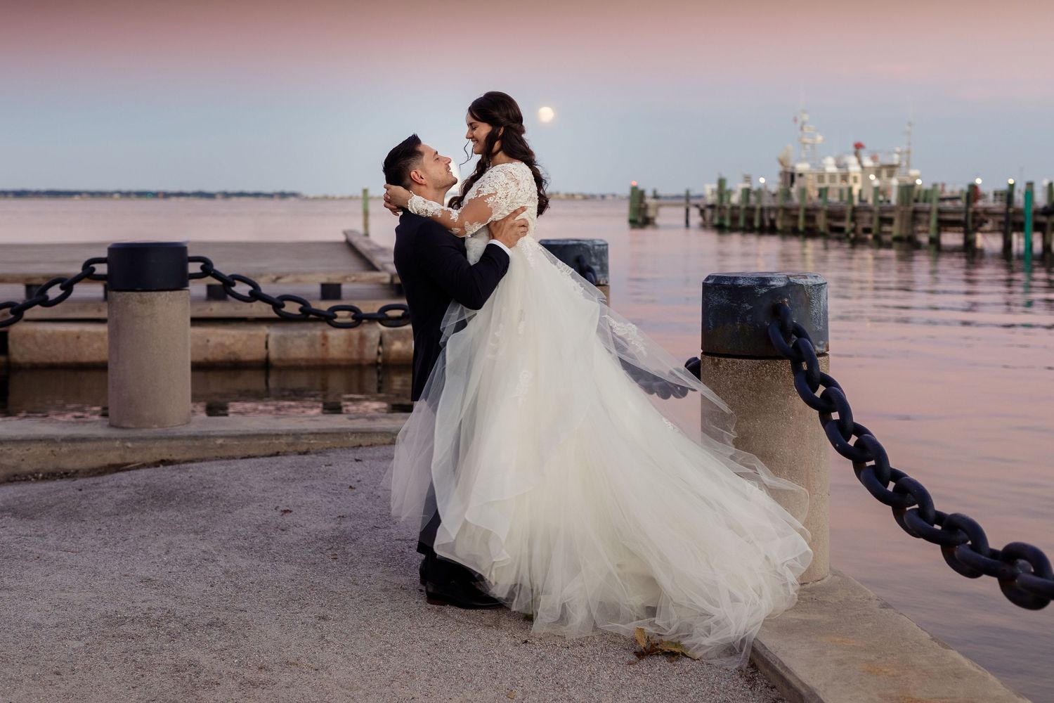 Couple in wedding attire dancing on waterfront pier at sunset.