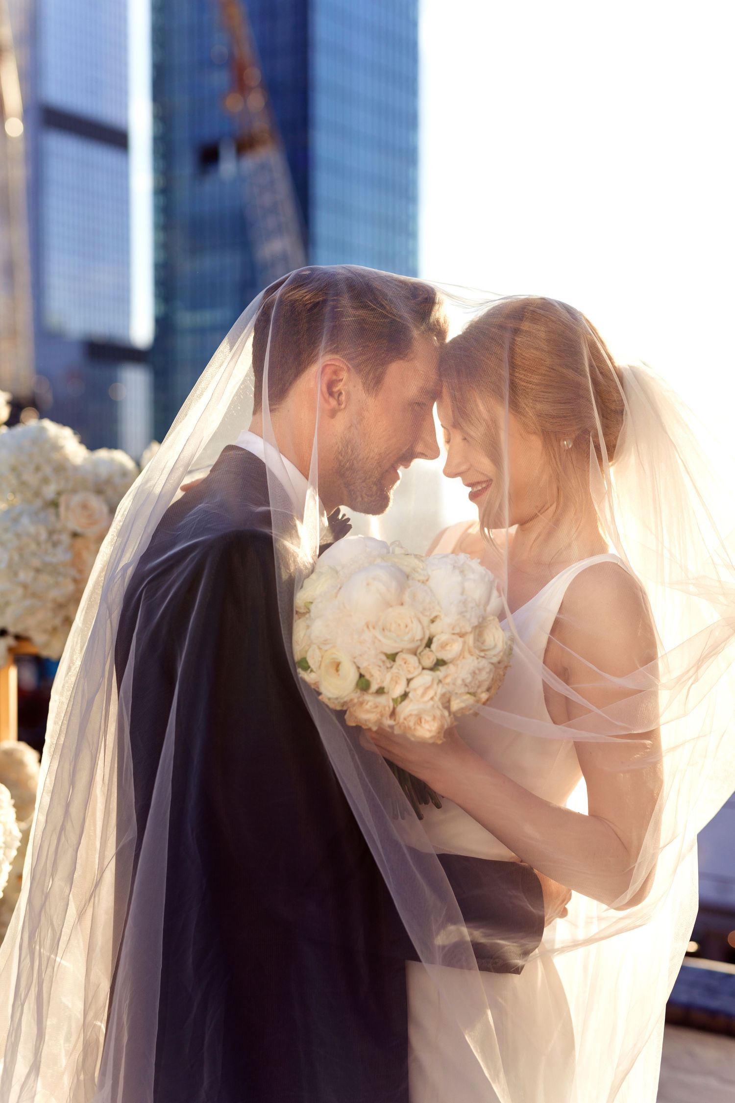 Sunlit couple with white bouquet and flowing veil against urban skyline.