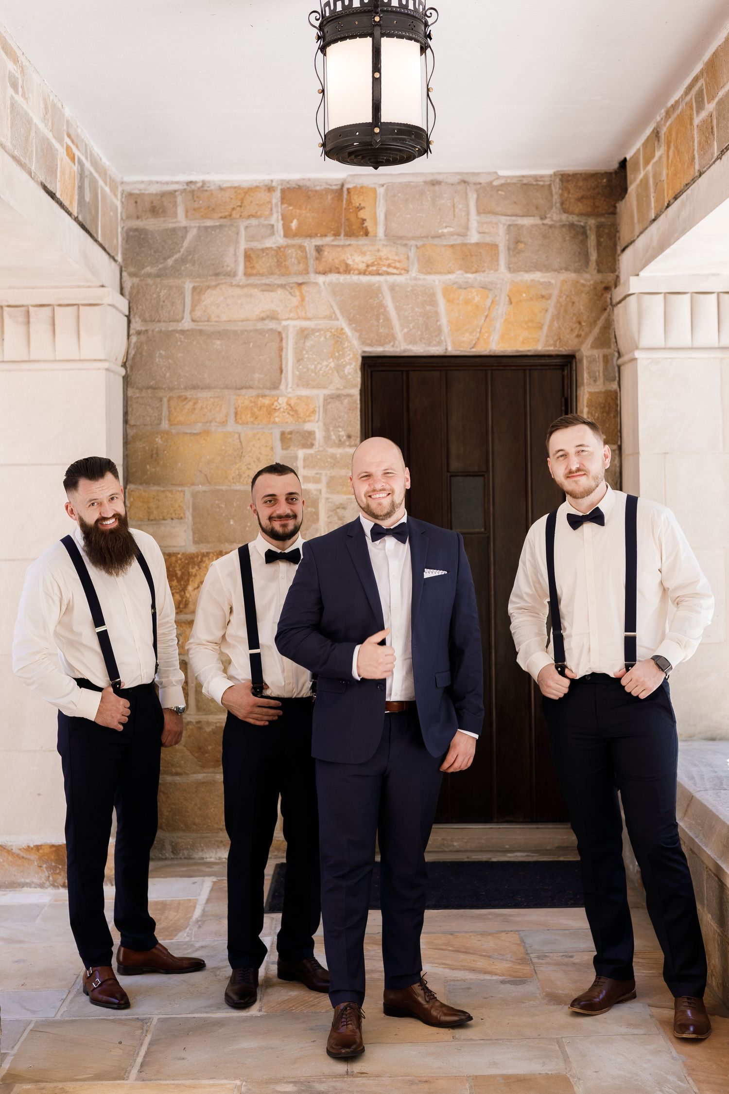 Groomsmen in suspenders and bowties pose together against stone wall interior.
