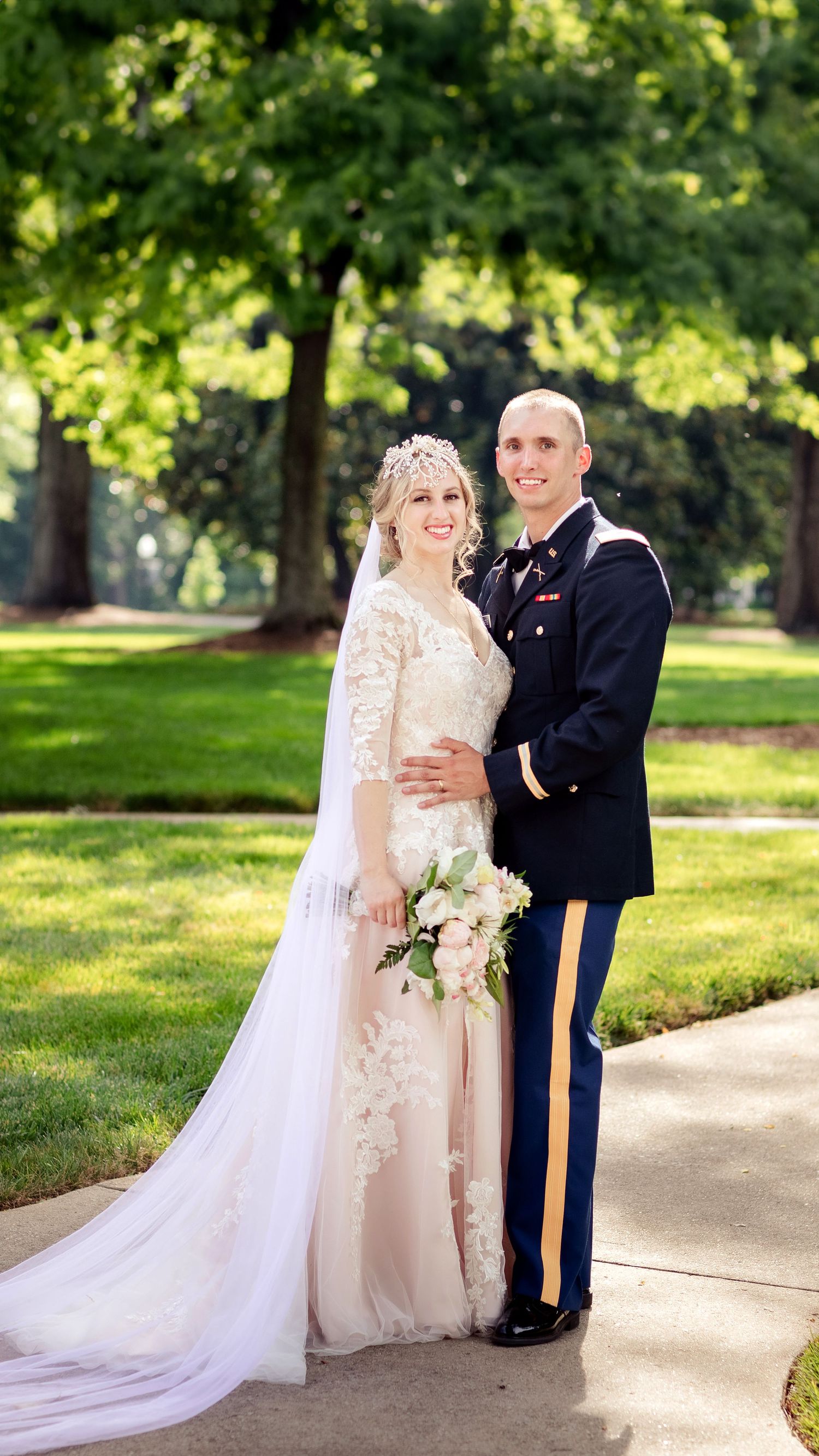 A military wedding couple poses outdoors in a park with lush greenery.