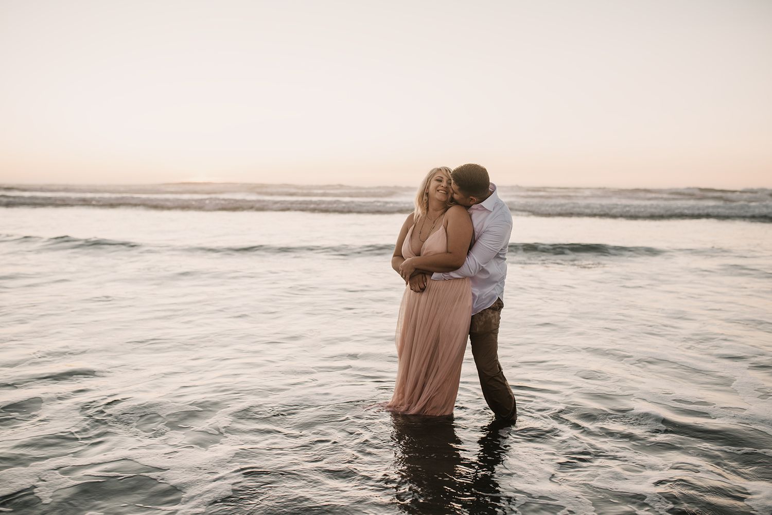 A couple standing in shallow ocean waves share an intimate moment at dusk.