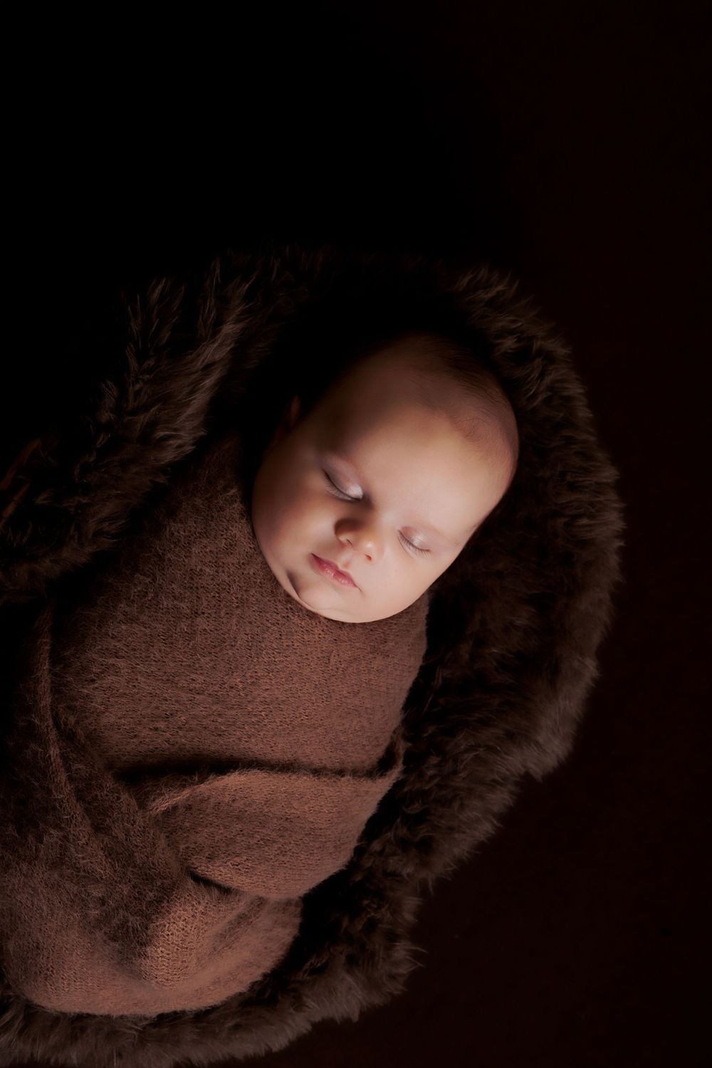 Newborn baby sleeping peacefully in brown swaddle with furry brown trim against dark backdrop.