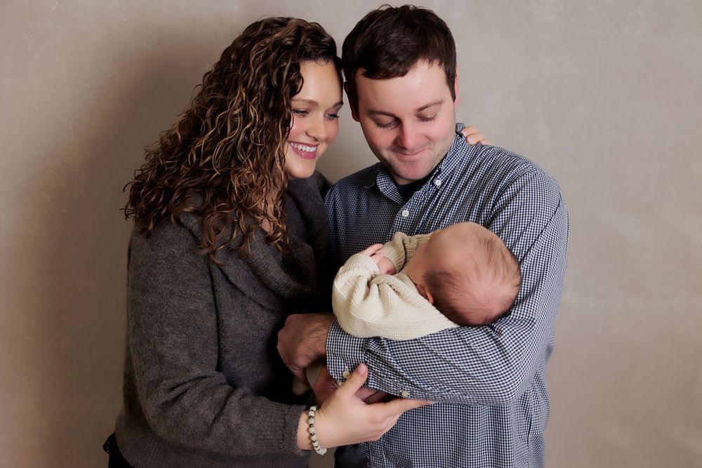 New parents sharing tender moment while holding sleeping newborn wrapped in cream blanket.