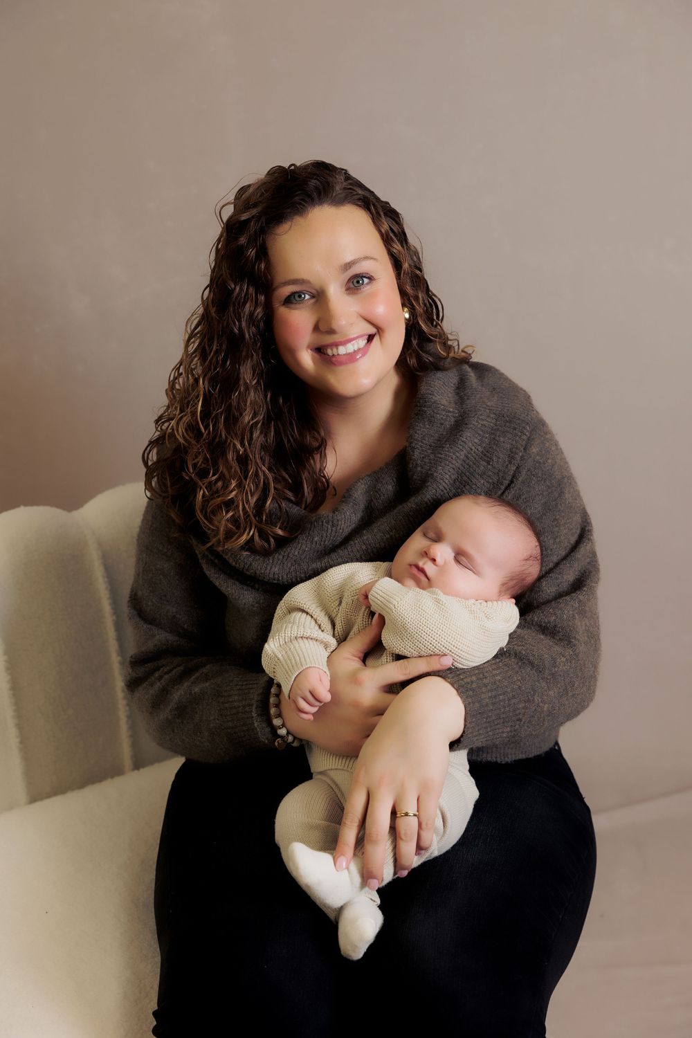 Person in grey sweater sits on cream chair holding sleeping infant.