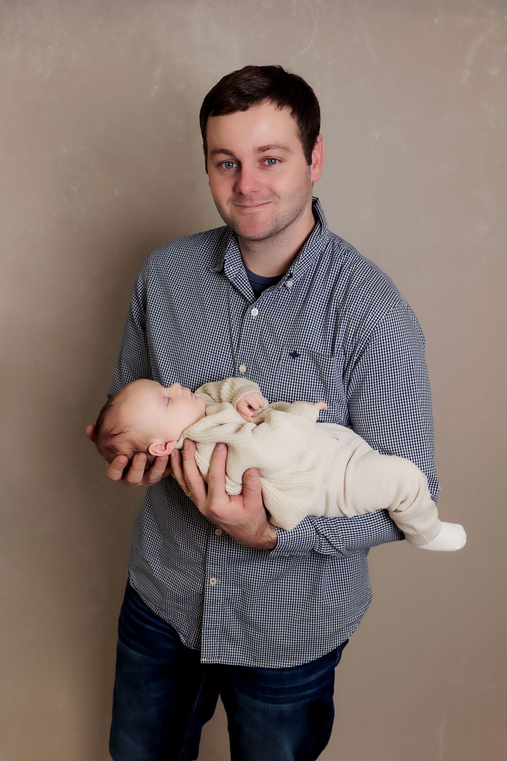 Parent in checkered shirt tenderly holding sleeping newborn in cream outfit.