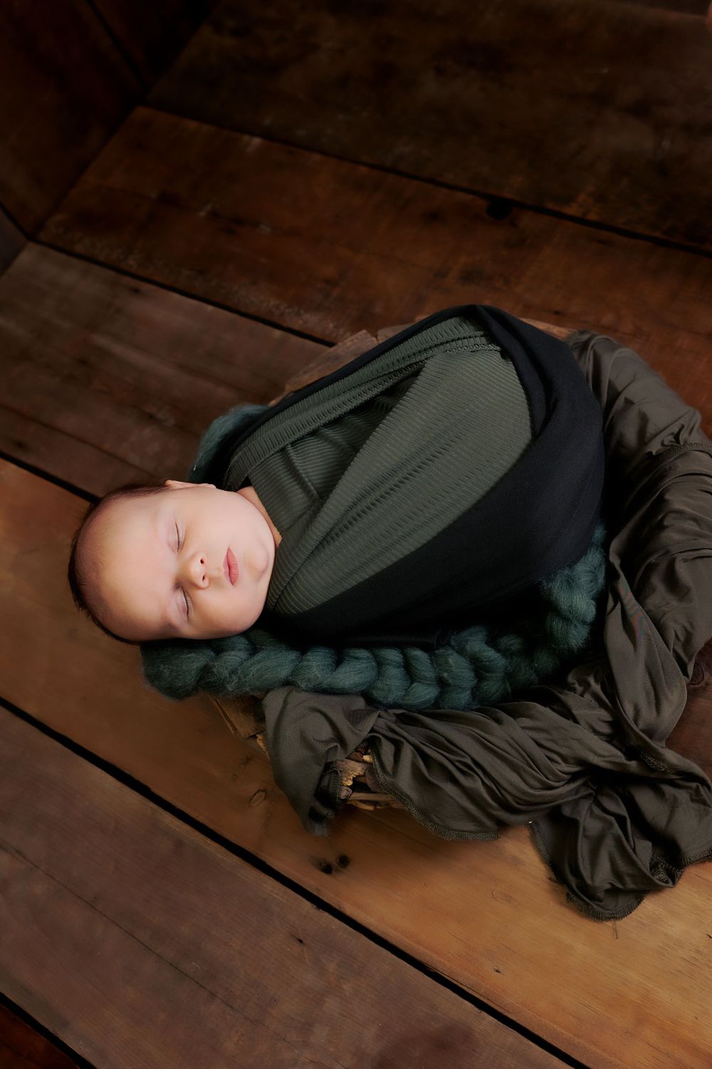 Sleeping baby wrapped in olive green swaddle on wooden floor.