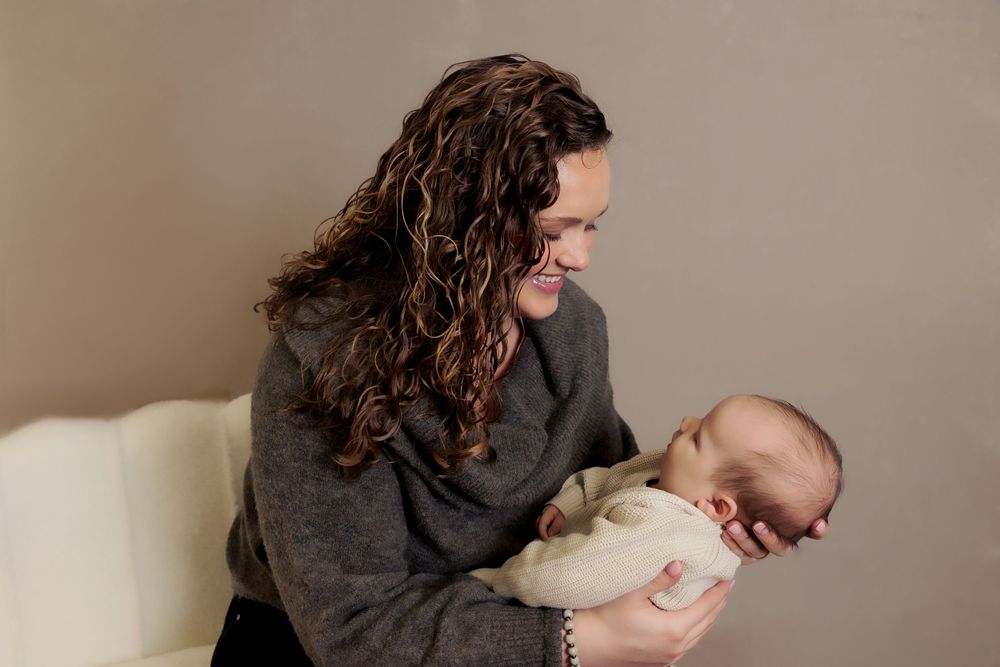 Person in grey sweater holding peaceful sleeping baby against neutral background.