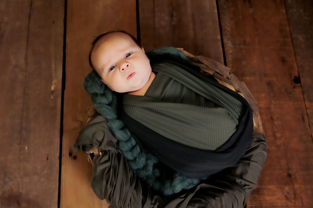 Baby resting peacefully in green and black swaddle on rustic wooden floor.