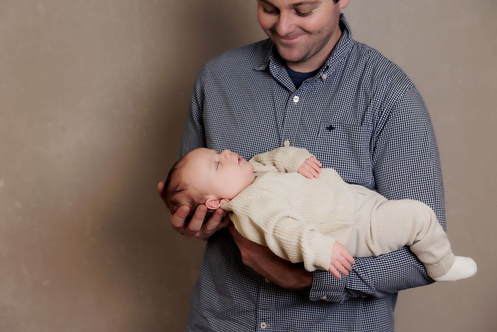 Adult in blue checkered shirt gazing lovingly at sleeping infant in arms.