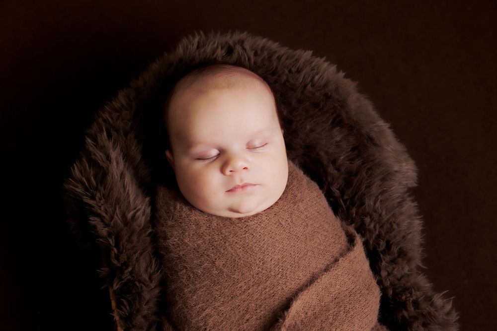Peaceful sleeping baby wrapped in brown knit against dark background with fur trim.