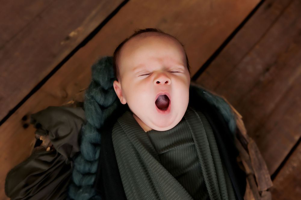 Yawning baby wrapped in olive green swaddle on dark wooden floorboards.