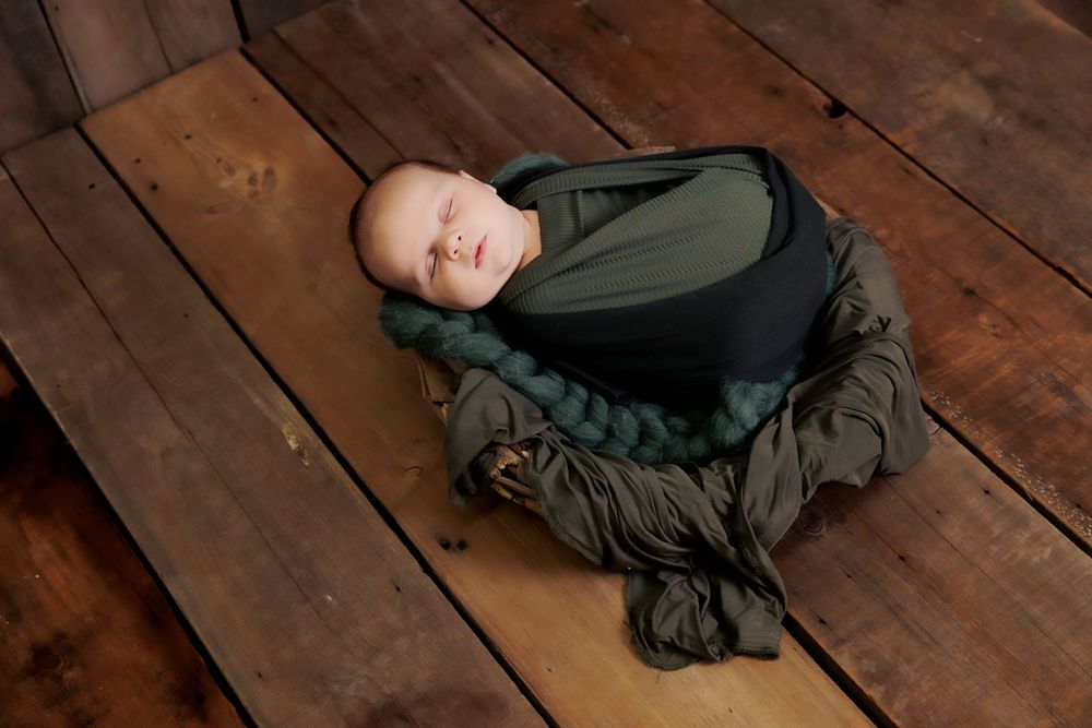 A newborn baby swaddled in olive and gray laying on wooden floorboards during a photography session.