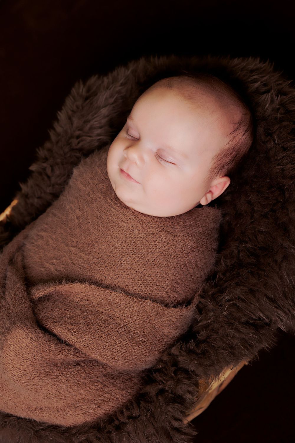 Sleeping infant wrapped in brown knit fabric on soft furry brown blanket.