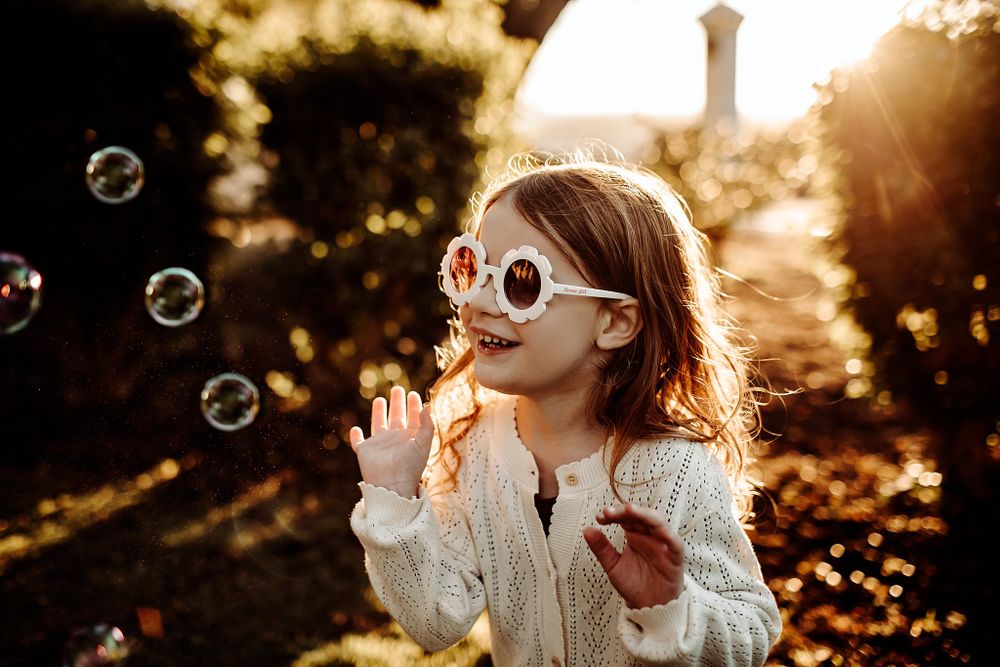 girl wears white sunglasses while bubbles float by during playful family session