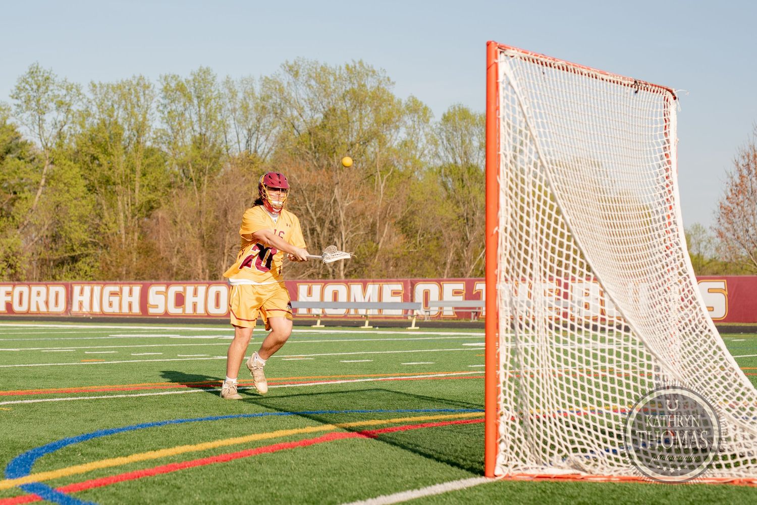 Lacrosse goalie in orange uniform stands ready to defend the goal on a turf field.