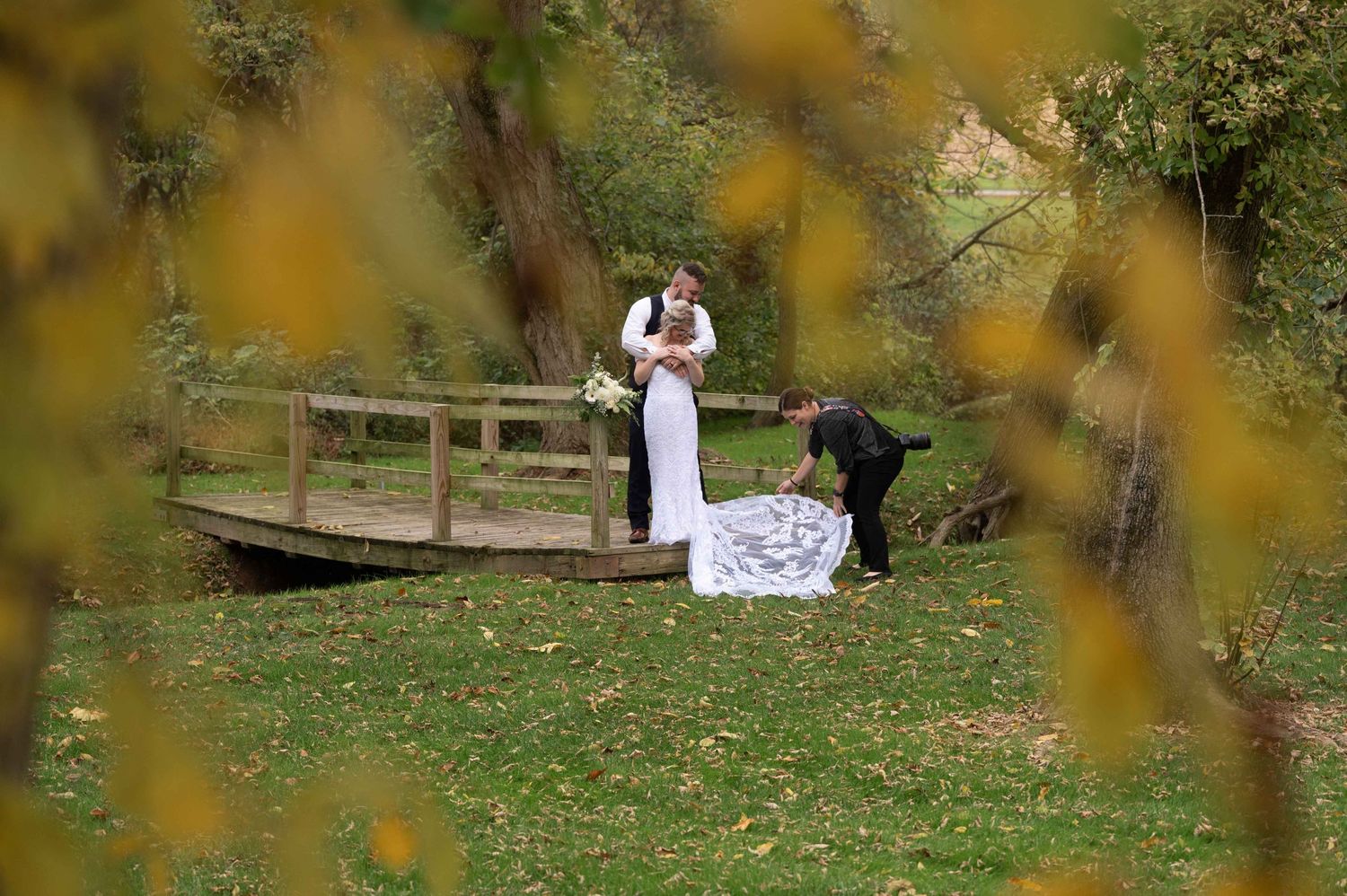 A photographer arranges a wedding dress train on a wooden bridge during an autumn photoshoot.