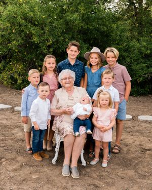 Elderly grandmother seated outdoors holding baby surrounded by eight grandchildren smiling.