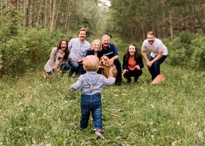 Toddler walking toward golden retriever dog with family watching and laughing in forest meadow.