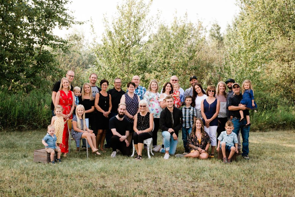 Large extended family of about 25 people poses together on a green lawn surrounded by lush trees in summer.