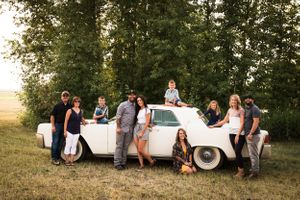 Extended family of ten poses around a classic white vintage car in a sunny green field at golden hour.