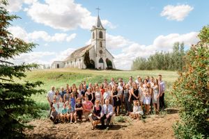 Large multigenerational family of about 60 people gathers in front of a historic white country church on a sunny day.
