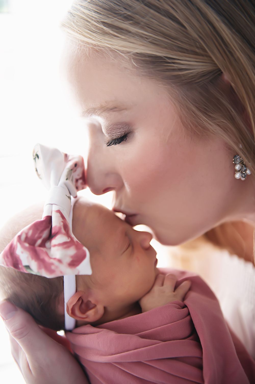 Mother gently kisses newborn baby wrapped in pink blanket in tender moment.