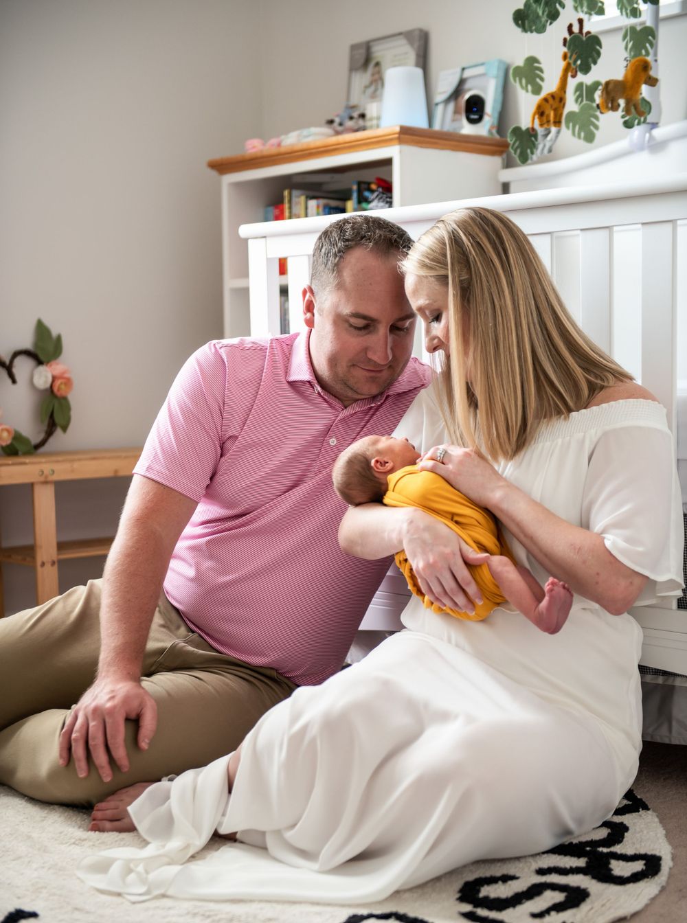 Parents cuddle with newborn baby in yellow onesie on white blanket.