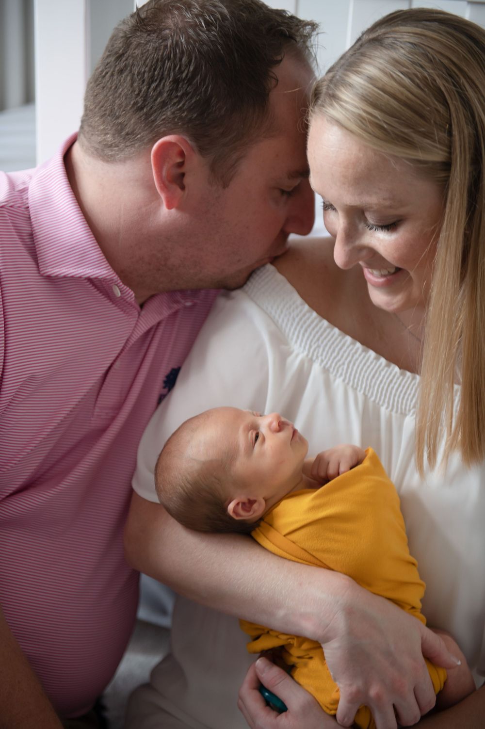 Intimate family moment with newborn baby in yellow onesie on white blanket.