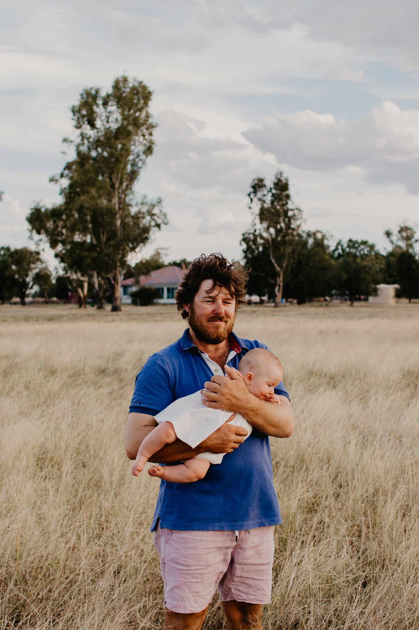 Dad, holding baby in grass paddock with their rural home in the background