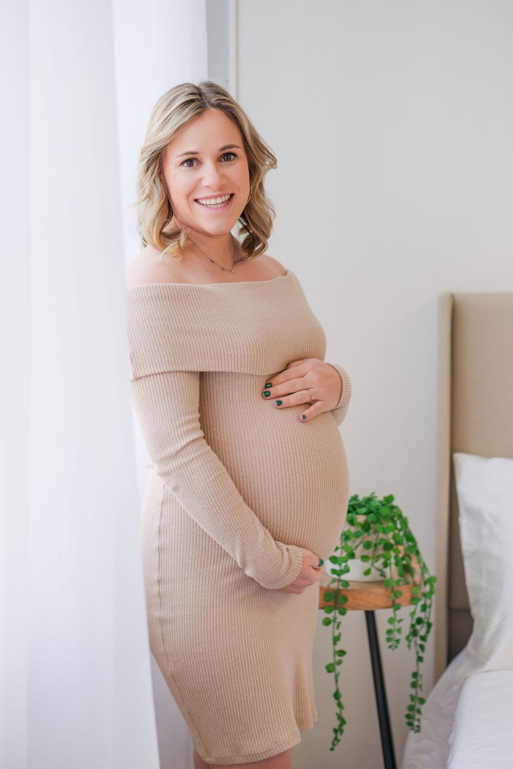 Expectant mother in beige ribbed dress smiling while cradling her baby bump in bright bedroom.
