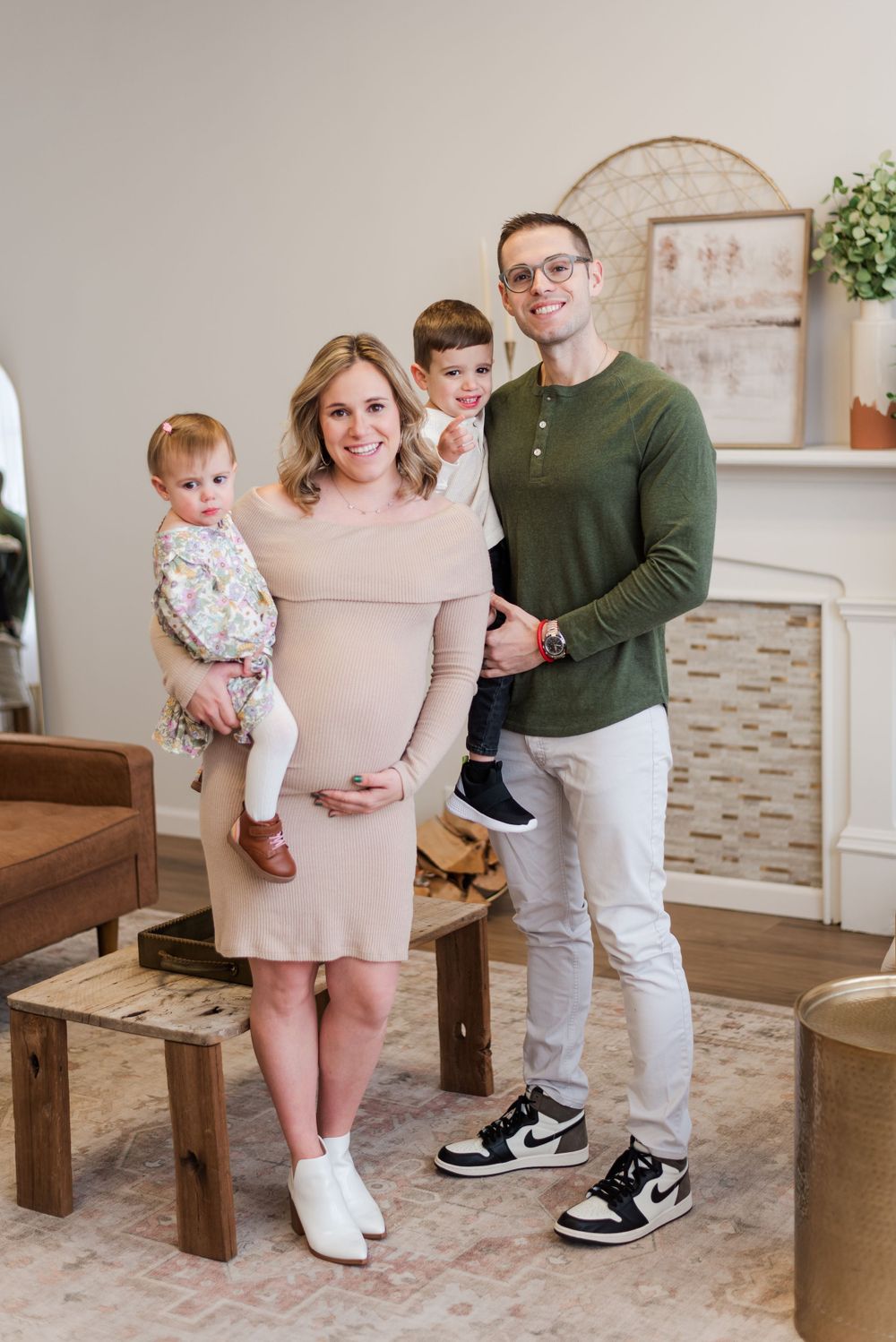Happy expecting couple wearing casual outfits stands with their small children in a cozy living room setting.