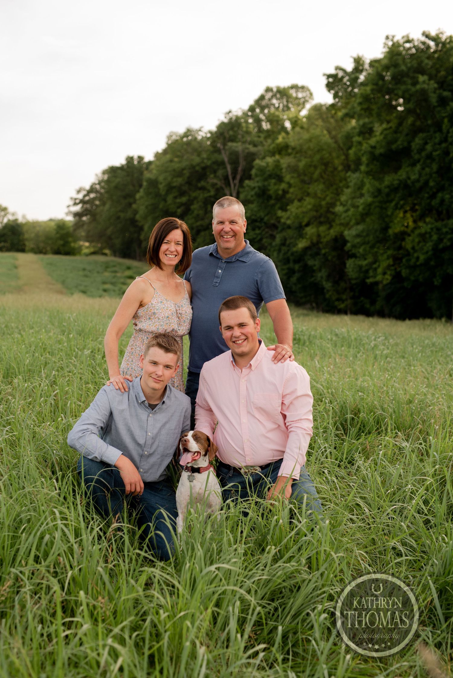 Family portrait with small dog in tall grass field during golden hour.