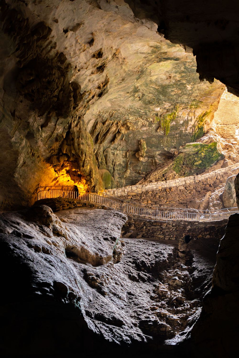 An illuminated cave pathway with rocky walls and a built-in walkway leading to a bright natural opening.