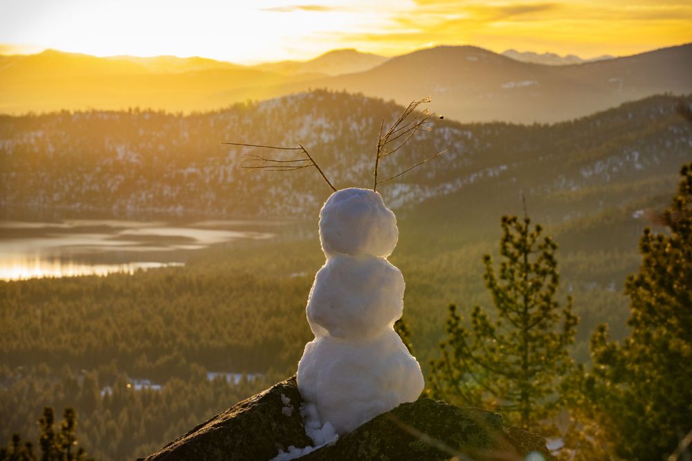 A snowman perched on a hilltop overlooks a stunning mountain landscape at golden sunset.