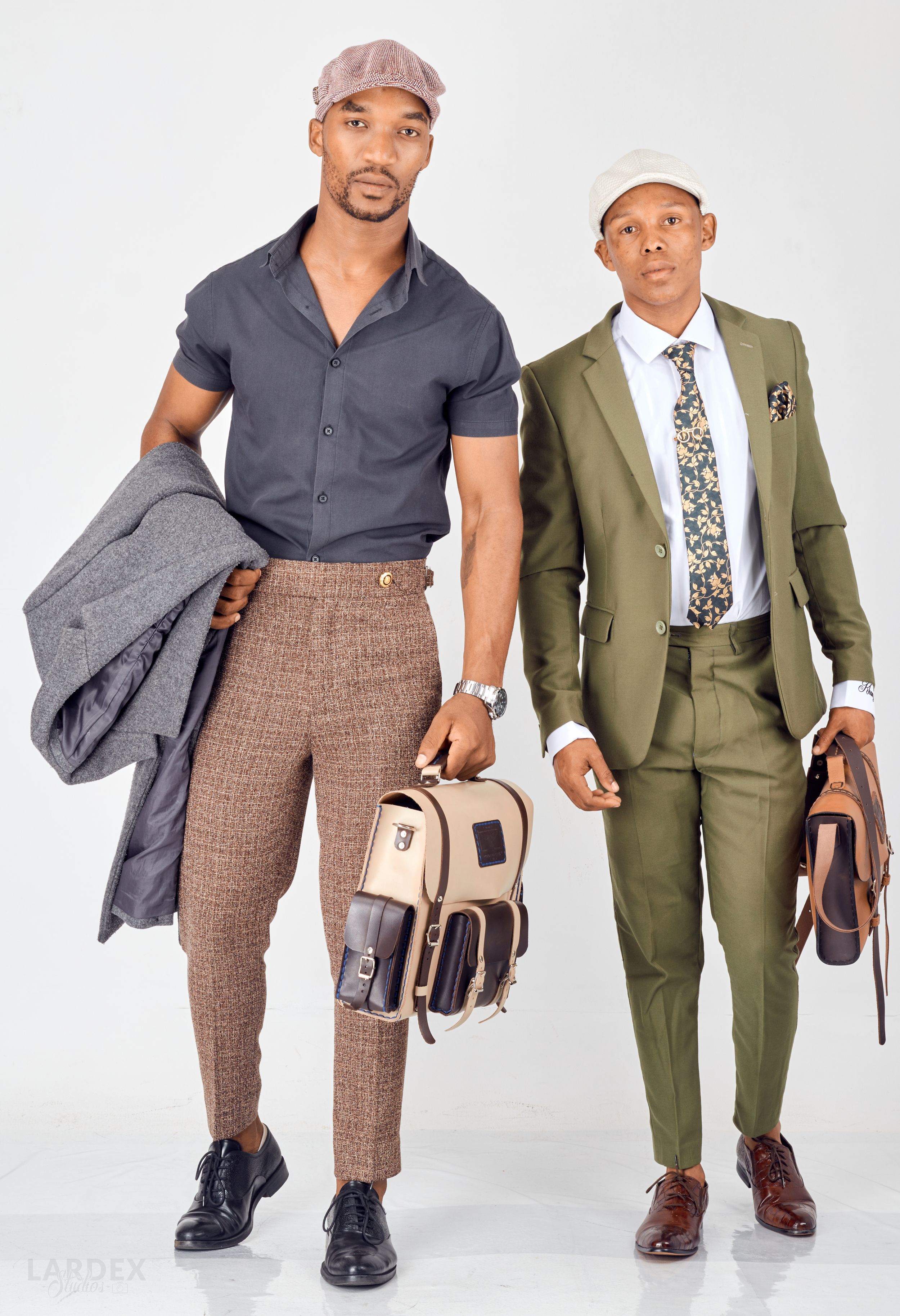 Two men in stylish outfits pose together, one in gray and brown casual wear and another in an olive green suit.