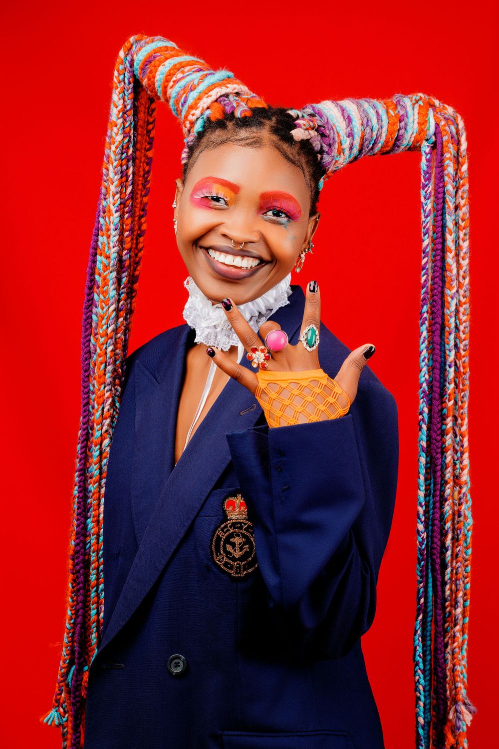 Playful portrait with colorful braided extensions and navy blazer against red background.