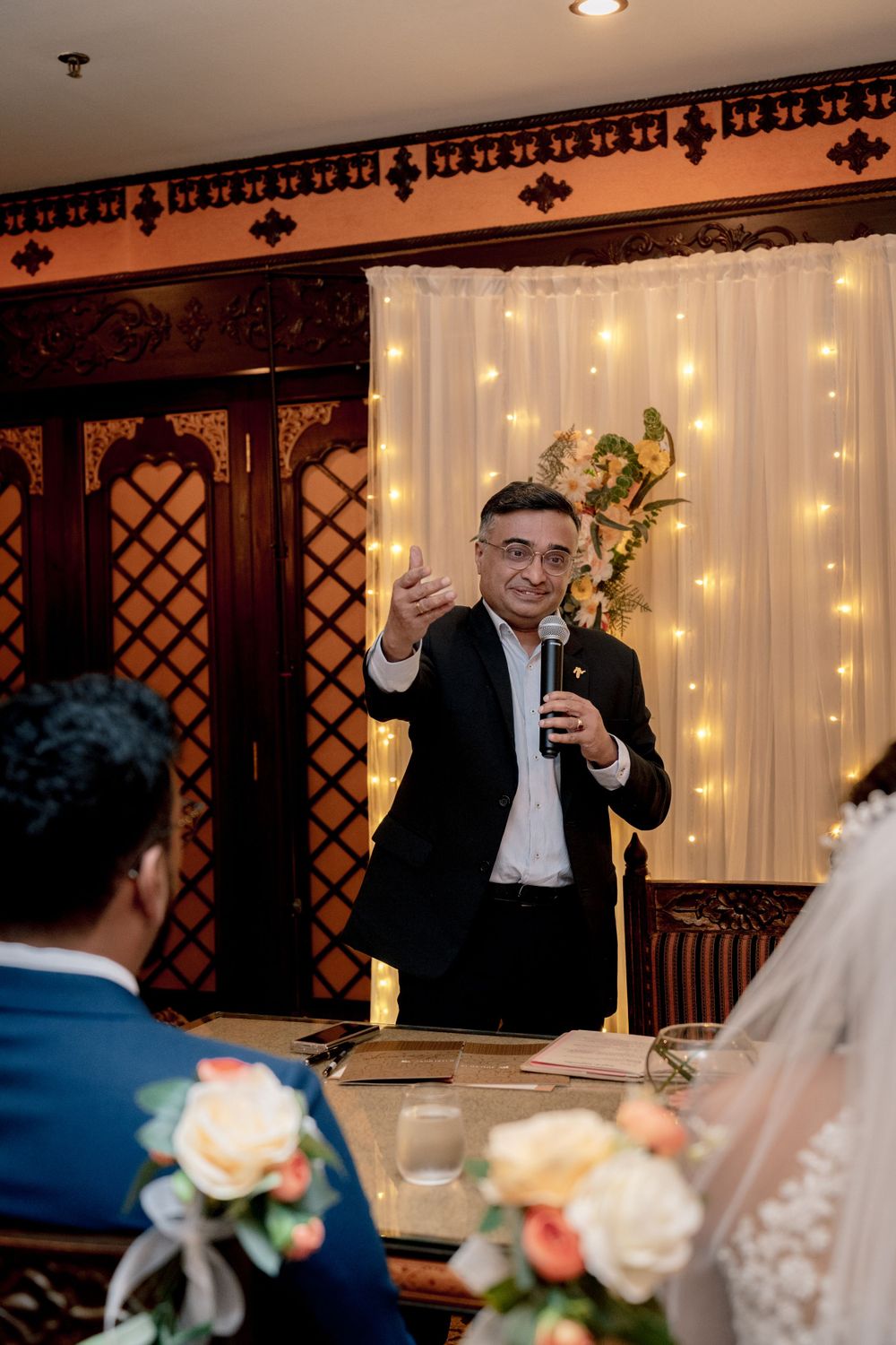 A person delivers a heartfelt speech with a microphone at a wedding reception against a backdrop of twinkling lights.