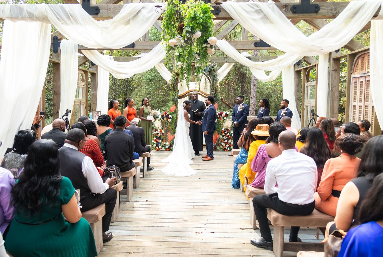Outdoor wedding ceremony setup with white drapes and wooden benches filled with guests.