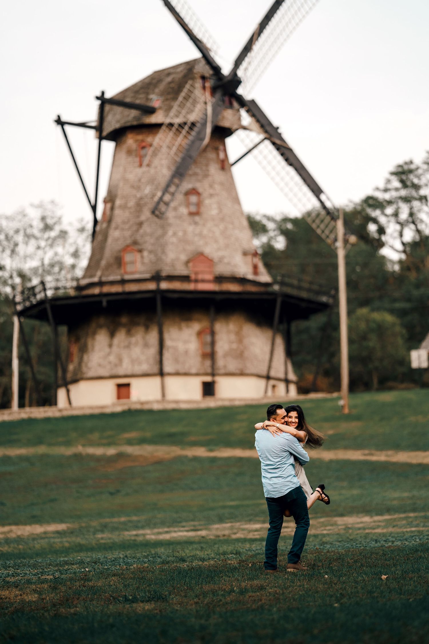 Man lifts woman, hugging amidst a green field with a windmill as backdrop, a moment of love and playfulness