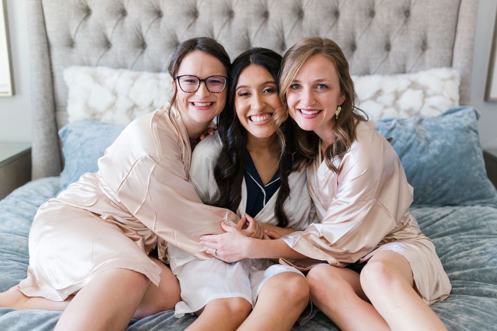 Three friends in robes share happy moment on bed during wedding morning preparations.