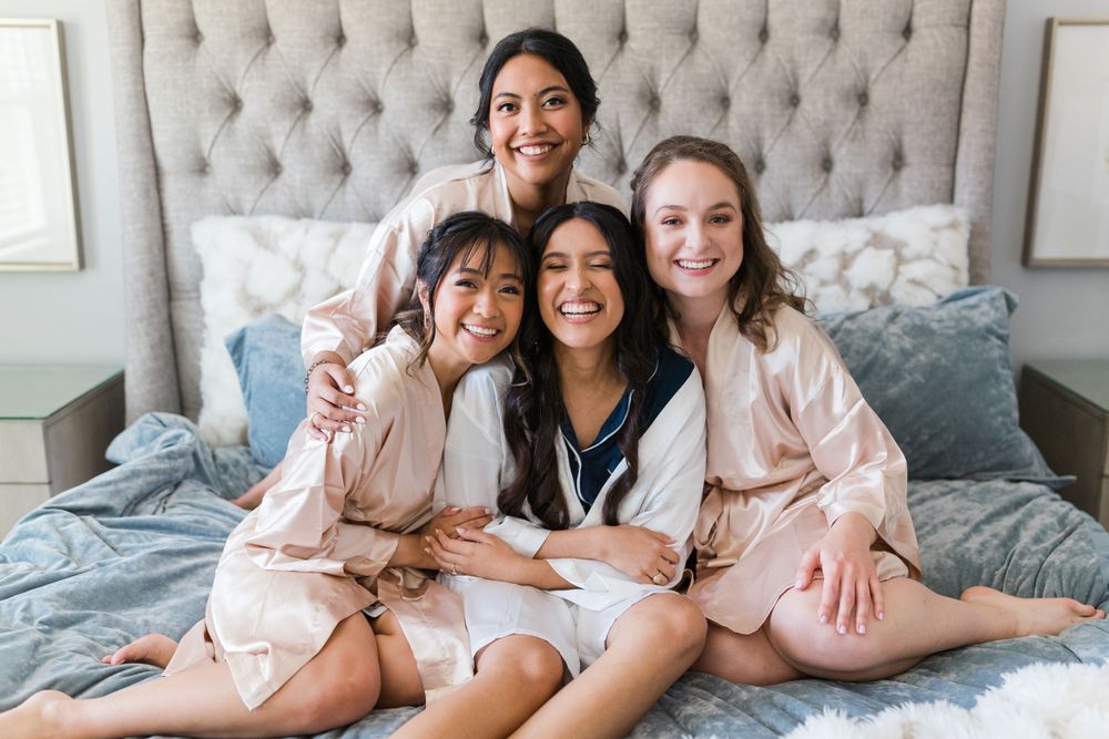 A group in matching pink silk robes sits together on a bed with a tufted headboard.