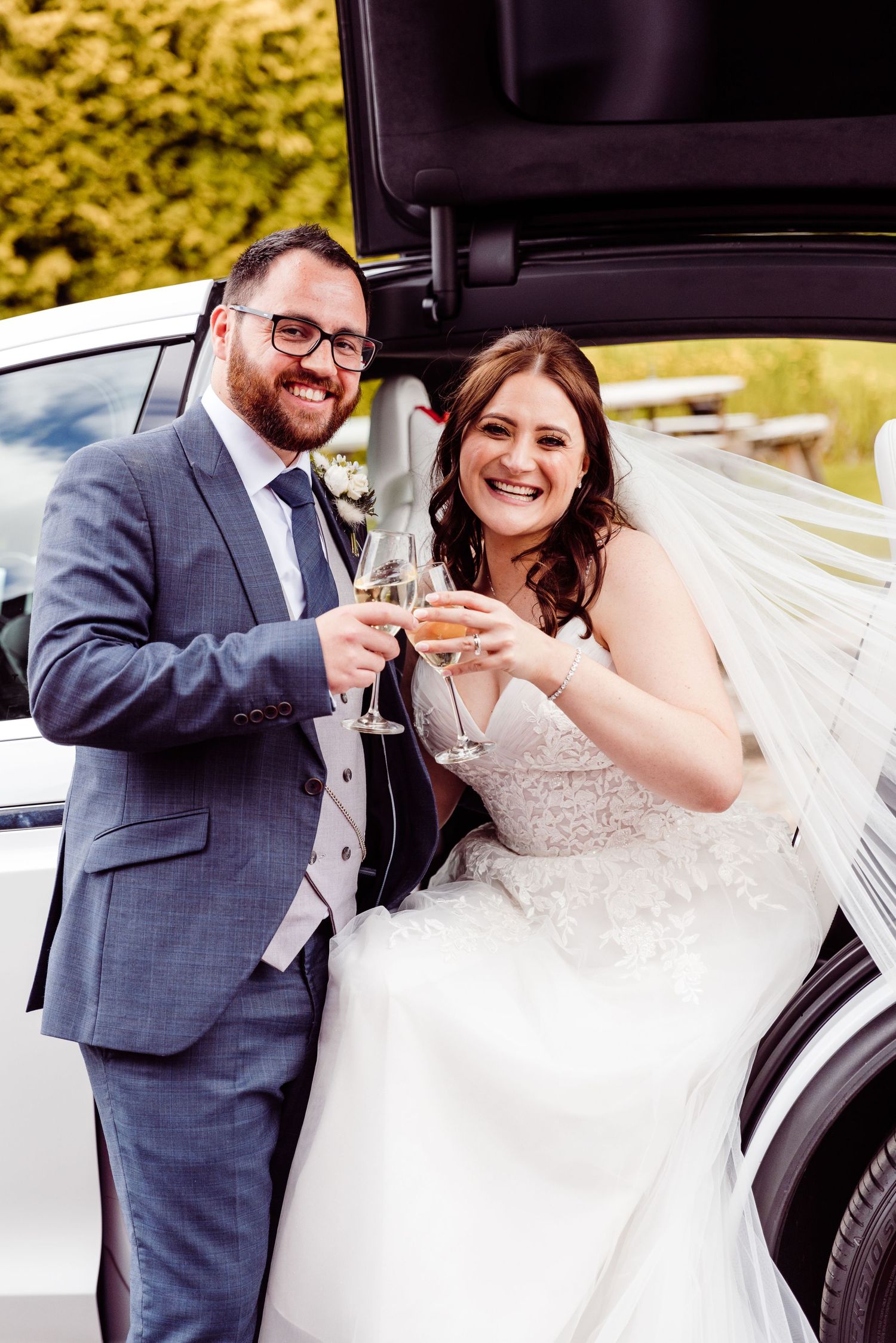 A wedding couple toasting with champagne glasses by their car.
