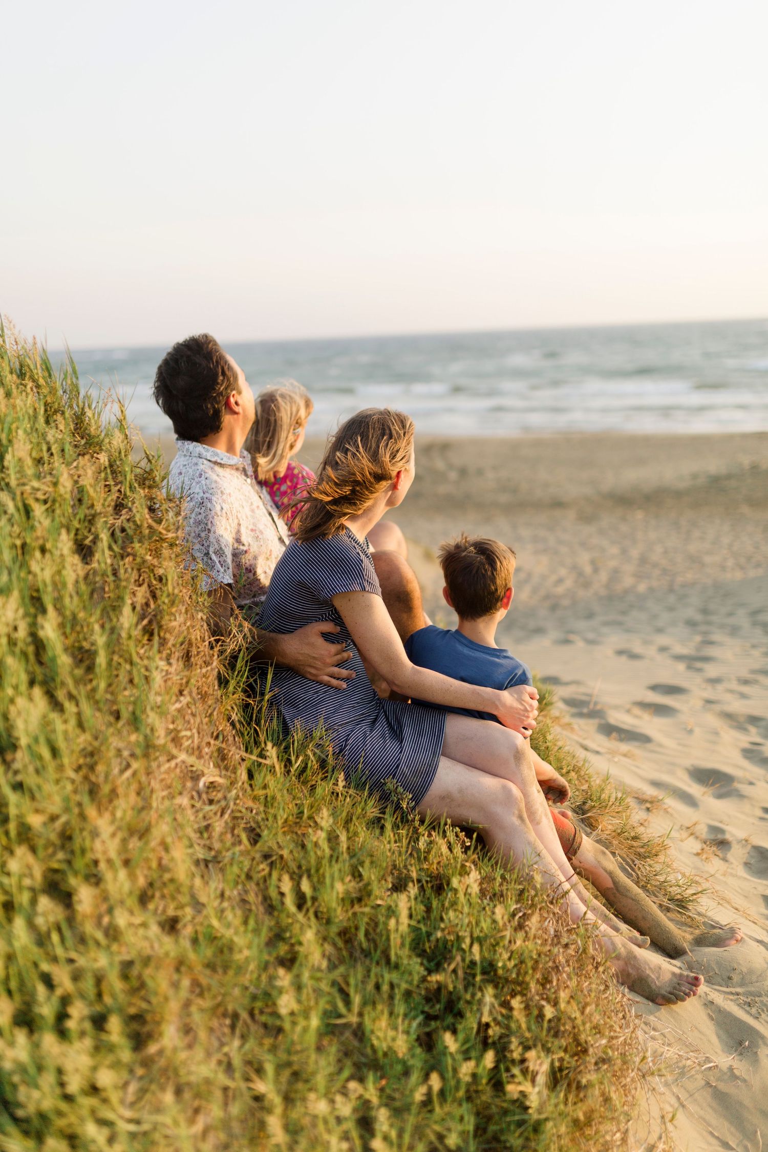Family sits on grassy dune overlooking beach at sunset.